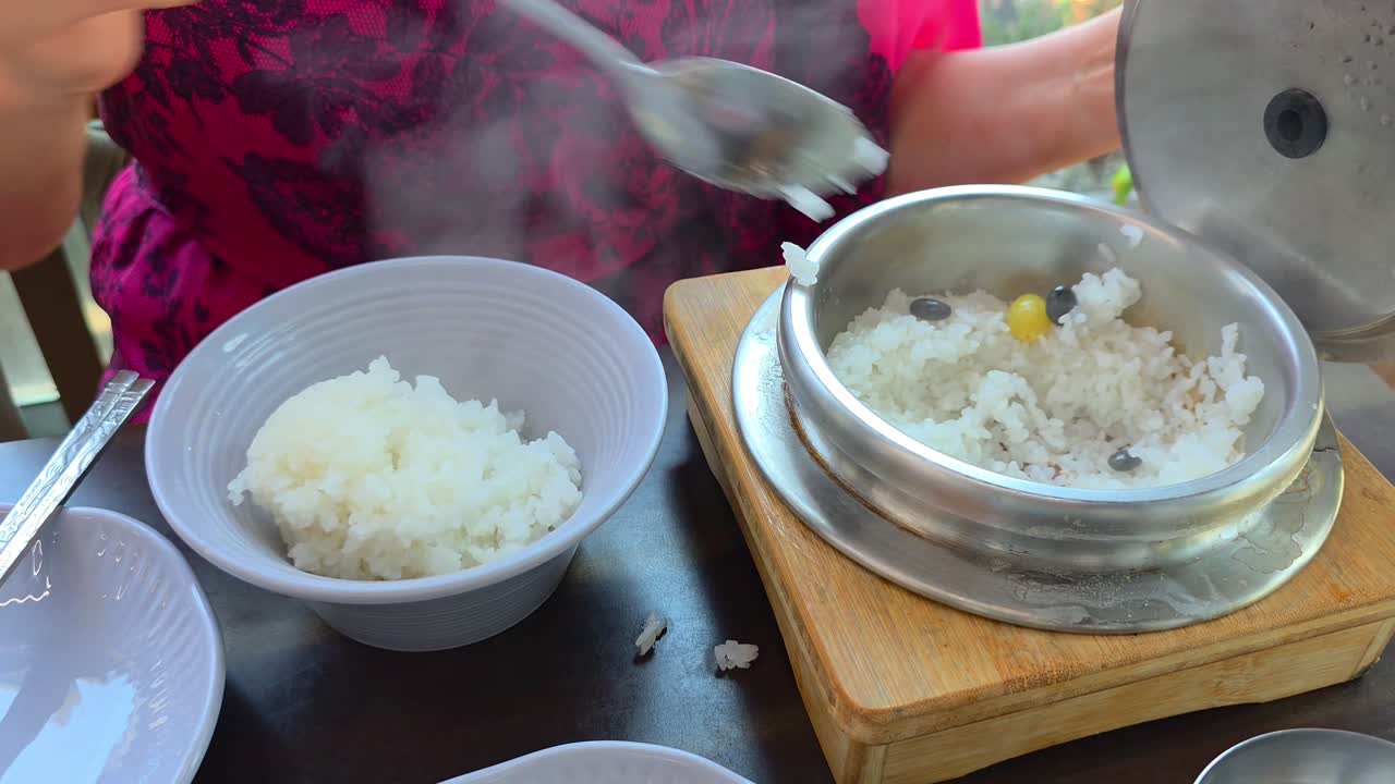 A close-up of a Korean woman traditionally eating rice by scooping hot rice from a metal bowl and pouring hot water to enjoy the remaining scorched rice