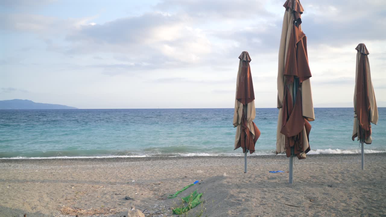 Coastal view of closed parasol sun umbrellas on empty sand beach at Kalamata, Greece. Wide view of Messinian bay on a quiet cloudy afternoon. Lest pan 4K footage