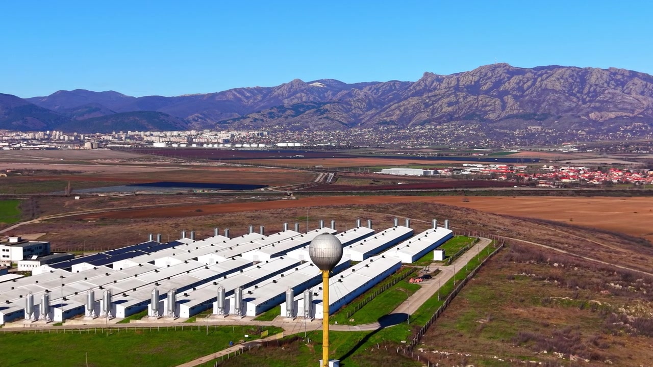 Complexo agrícola industrial com torre de água. Vista elevada de drone de edifícios paralelos brancos para gado, torre amarela com esfera, campos marrons e paisagem montanhosa sob céu azul claro.