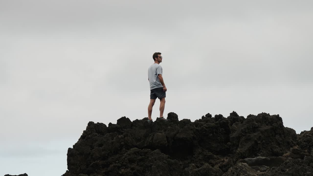 A man stands on jagged volcanic rocks under an overcast sky, gazing into the distance along the wild, dramatic coastline of Madeira, Portugal.