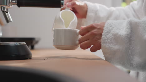 woman adds cream to coffee at table closeup. relaxed lady pours milk into cup from jug in sunny kitchen. preparing delicious caffeine drink in morning