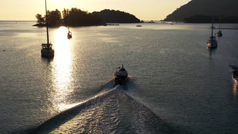 cinematic aerial drone following behind on a fishing boat out on the andaman sea at sunset with beautiful golden sun shimmering reflection on water surface capturing yachts and island silhouettes