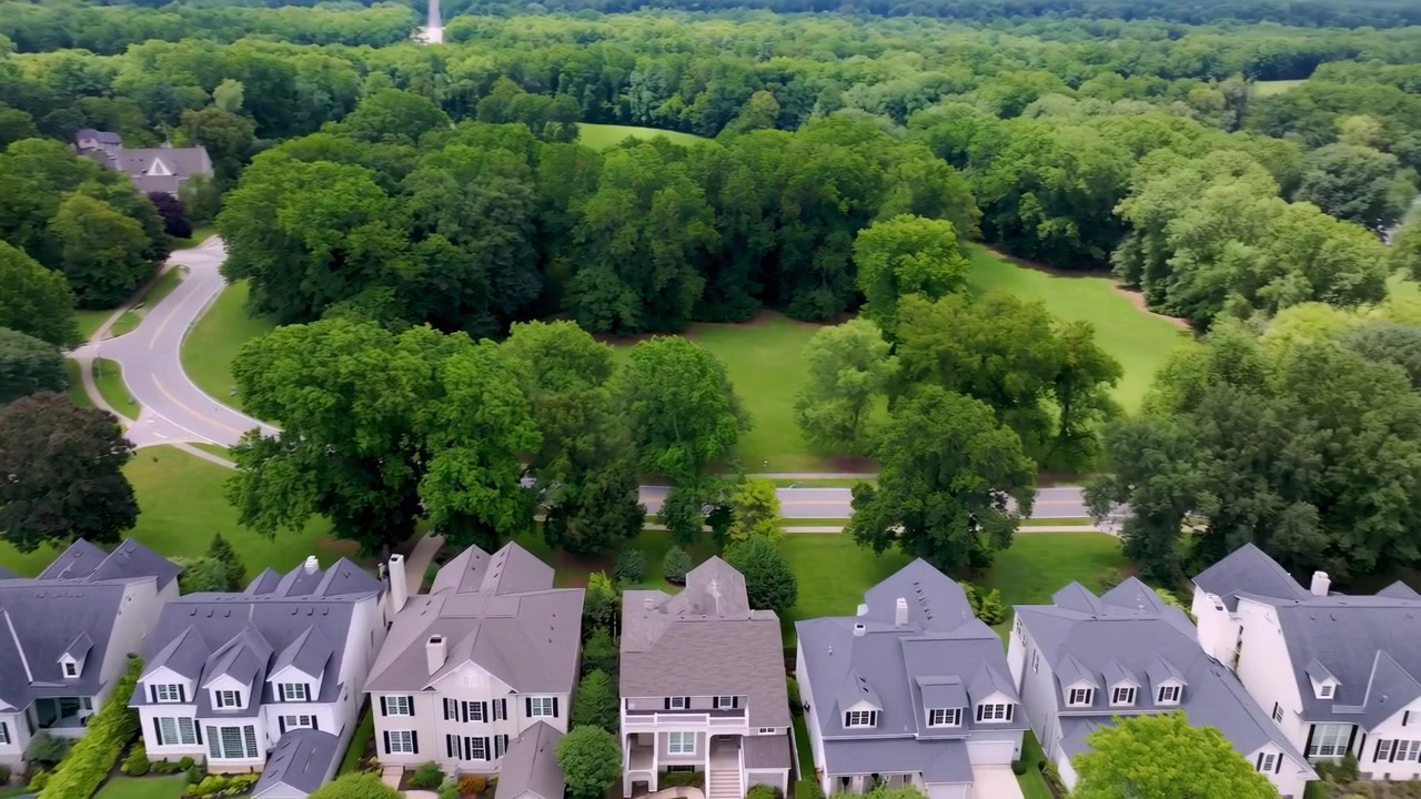Elevated oblique view showing row of upscale detached homes beside curved road, with pitched roofs, dormer windows, chimneys, front porches, driveways, clipped hedges, and surrounding deciduous