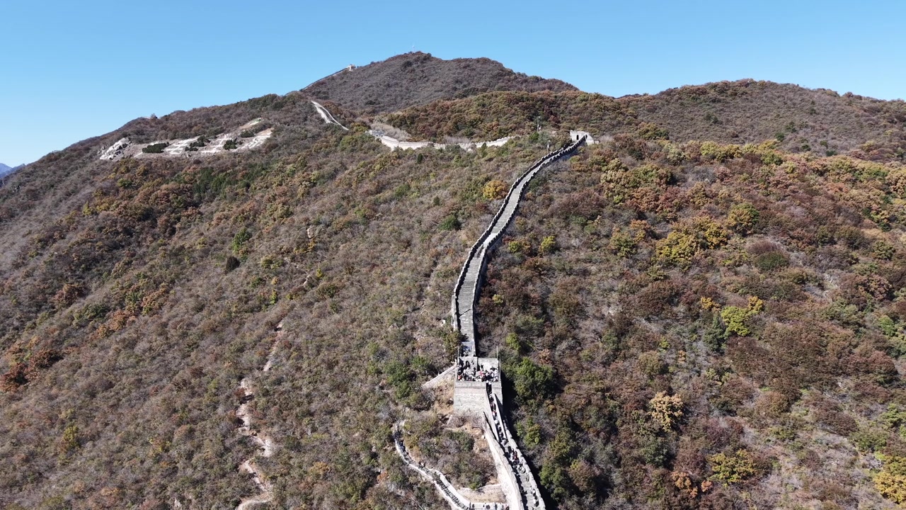 Vista aérea estática da Grande Muralha subindo diretamente em direção a um cume de montanha, um pico arredondado proeminente elevando-se acima da crista com encostas cobertas de outono abaixo, Distrito de Huairou, Pequim, China