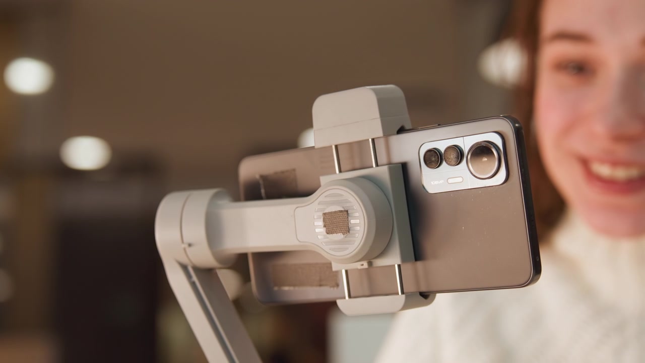 Close-up de smartphone preso em um grampo de gimbal de mesa, angulado em direção a uma pessoa sorridente ao fundo, com junta de ajuste visível, módulo de câmera traseira, luz quente de lâmpada, desfoque suave e mesa interna.