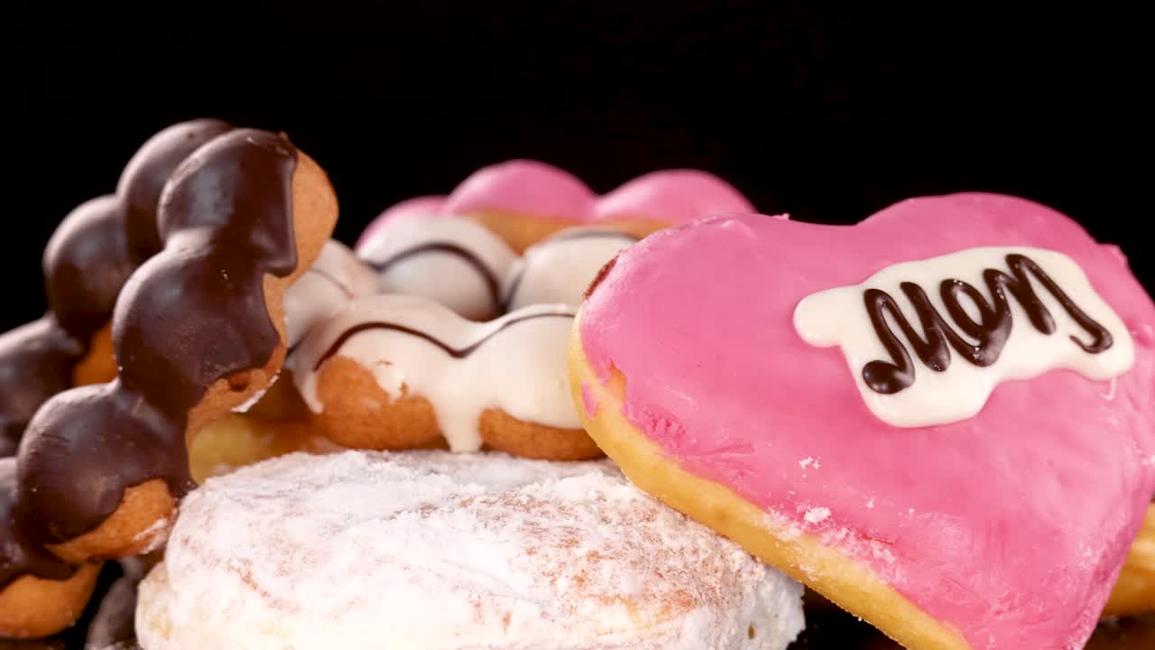 A close-up sequence of assorted donuts, including a heart-shaped pink iced donut with 'Mom' written on it, arranged on a black background with studio lighting and smooth camera movement