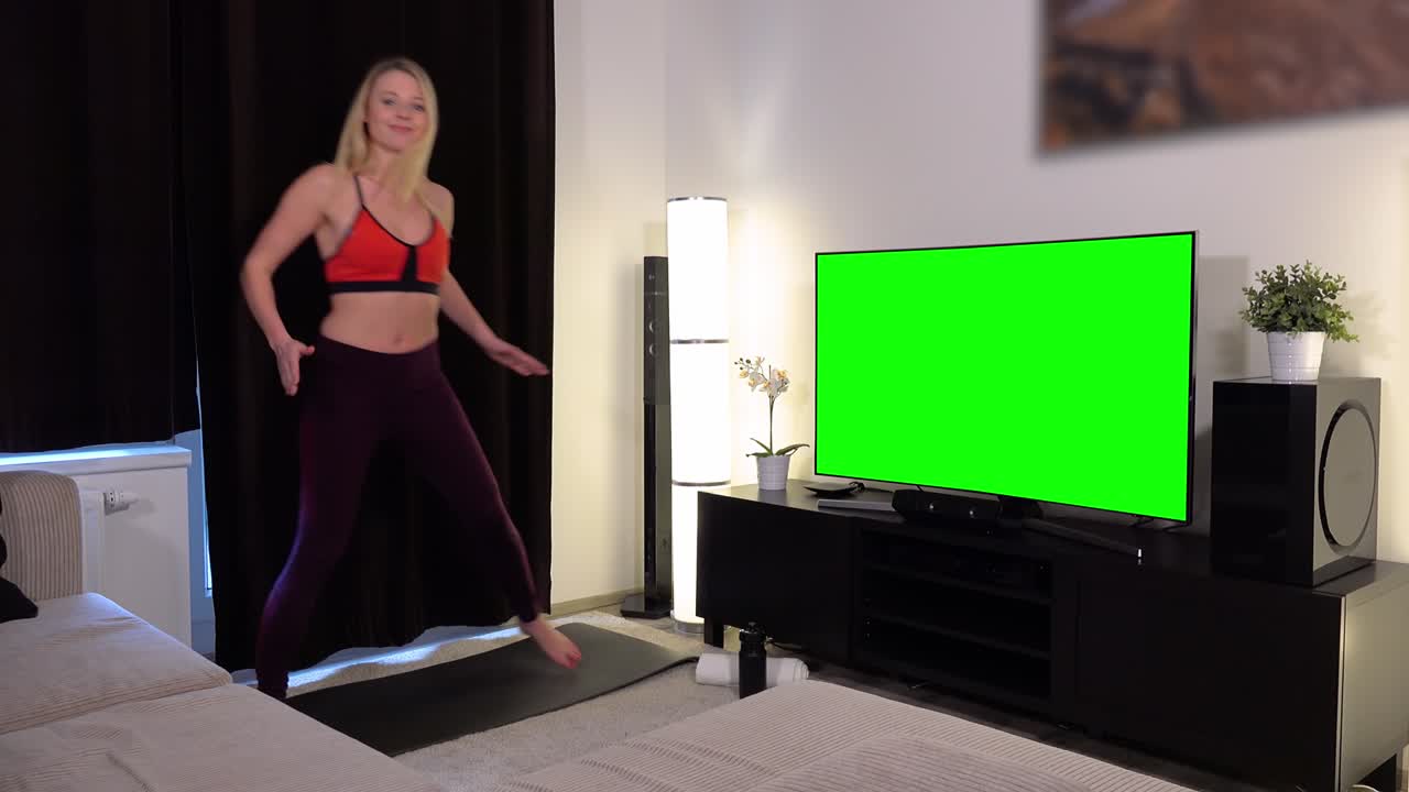 A young beautiful woman does a fitness dance in front of a TV with a green screen in an apartment and smiles at the camera