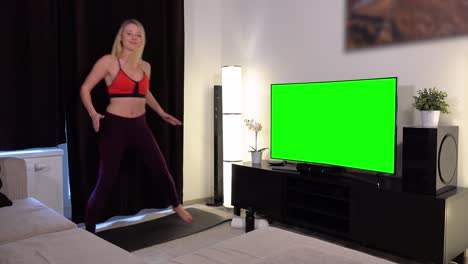 a young beautiful woman does a fitness dance in front of a tv with a green screen in an apartment and smiles at the camera