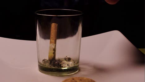 dark moments of depression and loneliness at home close up of a glass that is being used as an ashtray persons hand smoking holding cigarette putting ash inside the cup in very dark environment