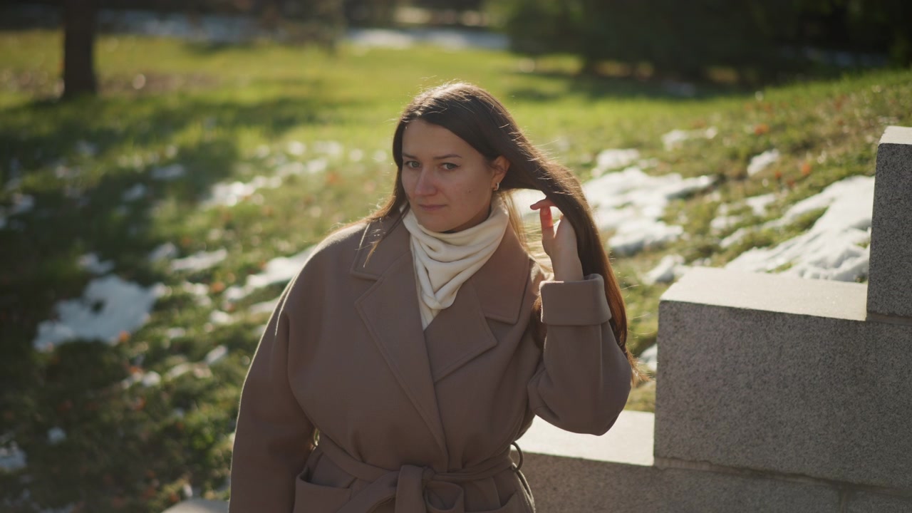 Primo piano di donna che si sistema i capelli e il cappotto in un ambiente di parco all'aperto con erba verde, chiazze di neve, alberi, illuminazione soffusa, espressione calma, dettaglio della texture, soggetto immobile. La donna indossa