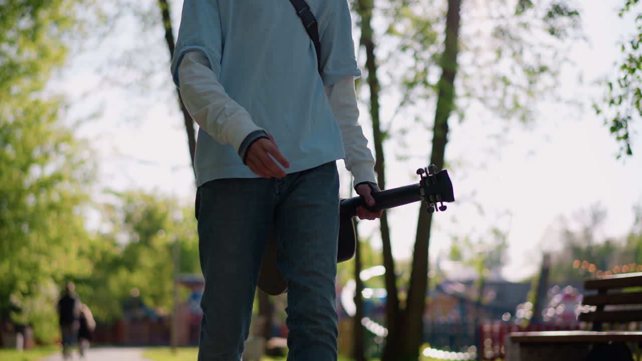 joven guitarrista con guitarra acústica caminando por un sendero arbolado; la luz matinal que se filtra entre las hojas lo ilumina a contraluz; viste vaqueros y sudadera informales con una correa para el hombro; camina relajado hacia un banco