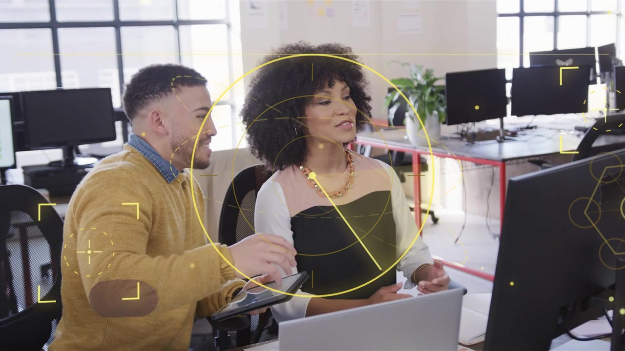 Leaning man mustard-sweater pointing at monitor at desk guiding woman reading file on tablet HUD