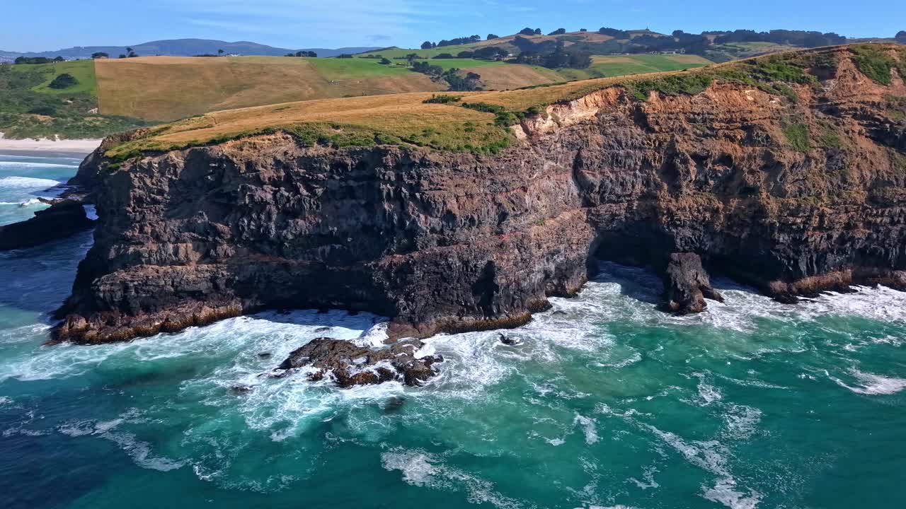 Drone side shot moving left to right along the cliffs of Smaills Beach, showing waves crashing on rocks, green vegetation, and a blue sky