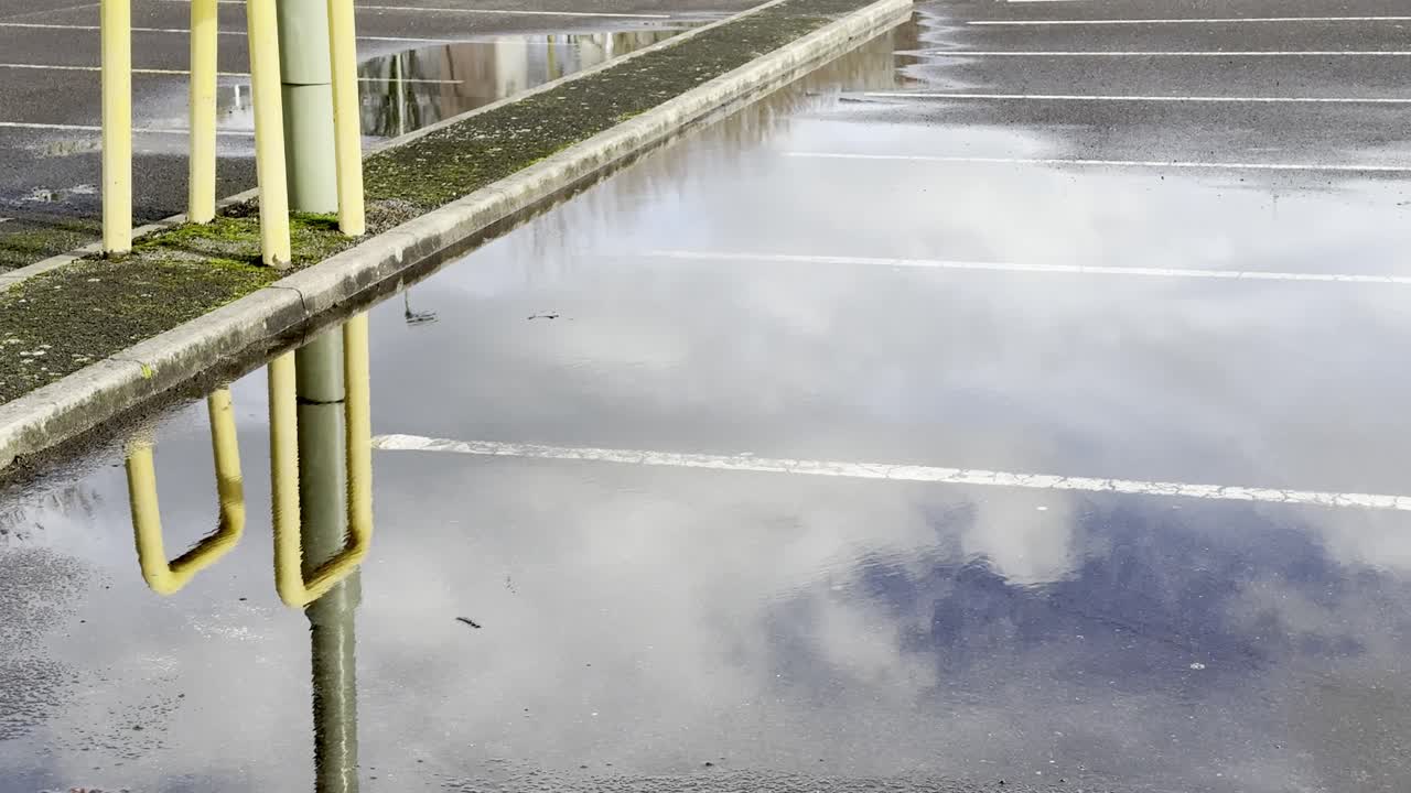 Mraky a letící pták odrážející se v dešťové kaluži na povrchu prázdného parkoviště.
