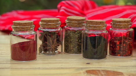 selection of jars with dried spices laid out in a row on wooden table with red cloth in the background