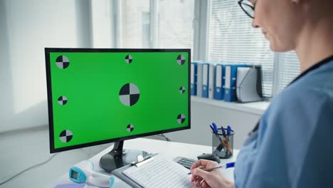 el médico consulta al paciente en línea a través de una computadora con pantalla verde, el clínico en uniforme azul habla y escribe en el clipboard en el lugar de trabajo en el hospital