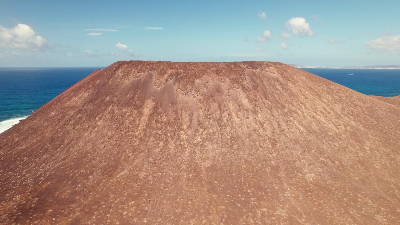 Aerial drone shot flying over ridge of Montaña de La Caldera on Lobos Island, Fuerteventura, highlighting the volcanic cone with the ocean and clear blue sky as a stunning backdrop.