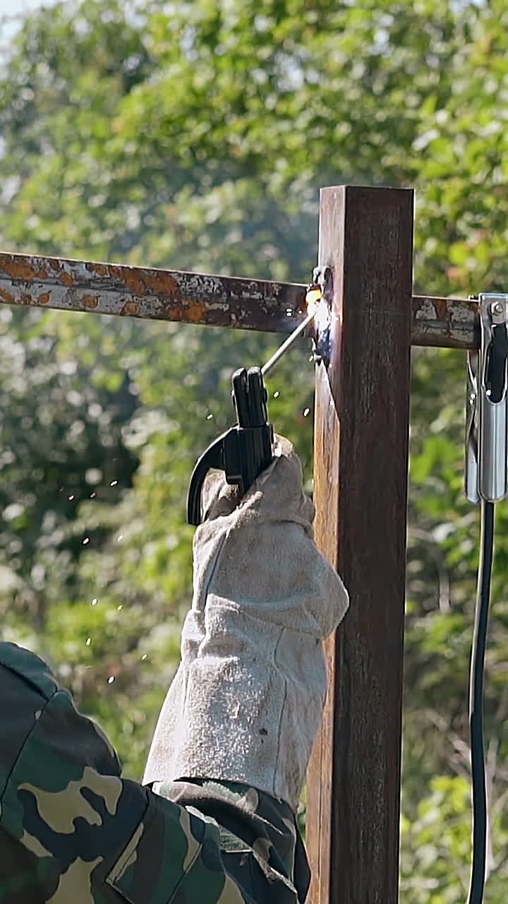 young man in protective gloves welds carcass of country house fence by trees slow motion backside view