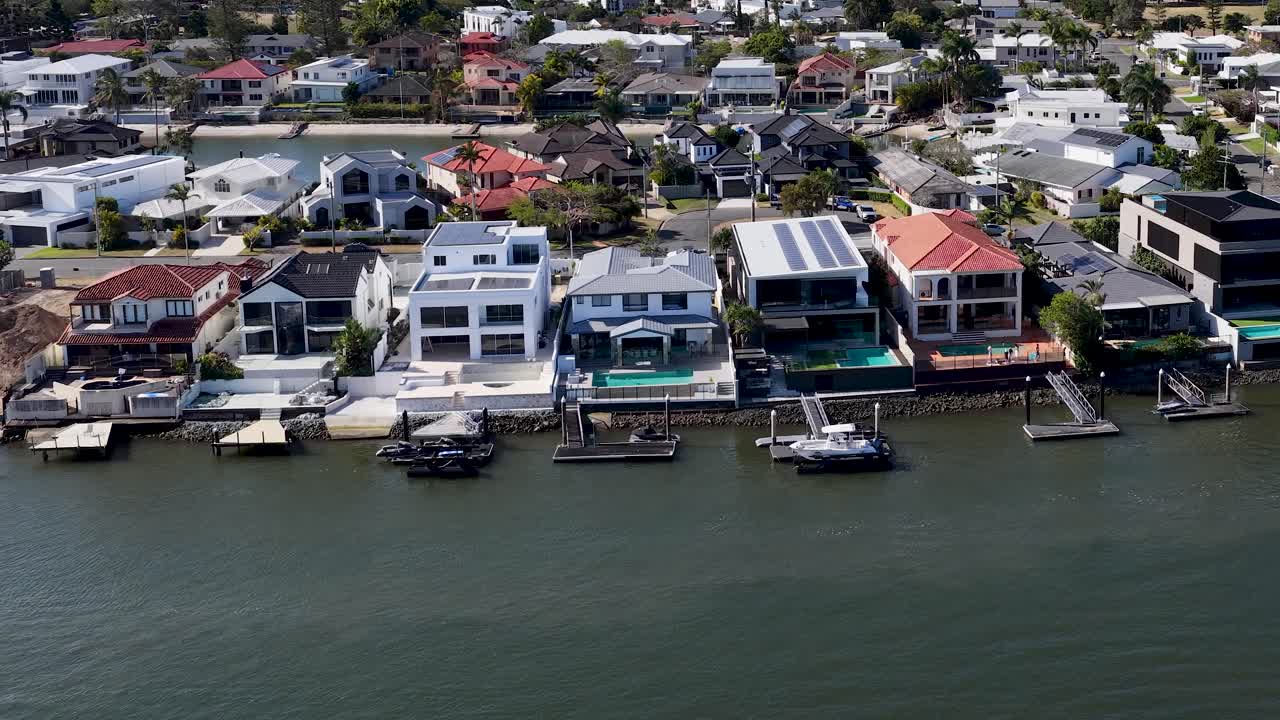 Drone glides above residential waterfront, modern houses, private docks, and calm water in daylight