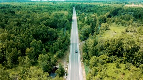 wide shot aerial drone footage tracking a sedan car cruising on an empty road surrounded by a beautiful large forest and river