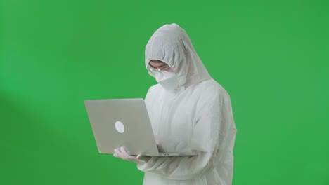 side view of asian man wearing personal protective equipment uniform ppe, medical face mask, holding and working with laptop computer in the green screen studio
