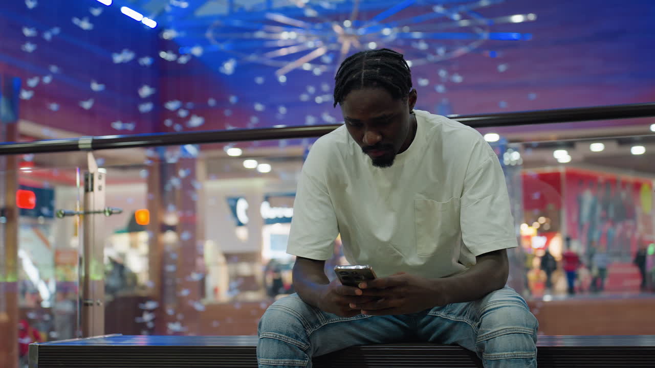 Celebrity man in white shirt sits alone on bench inside bright shopping mall, focused on phone while colorful lights and blurred shoppers in background