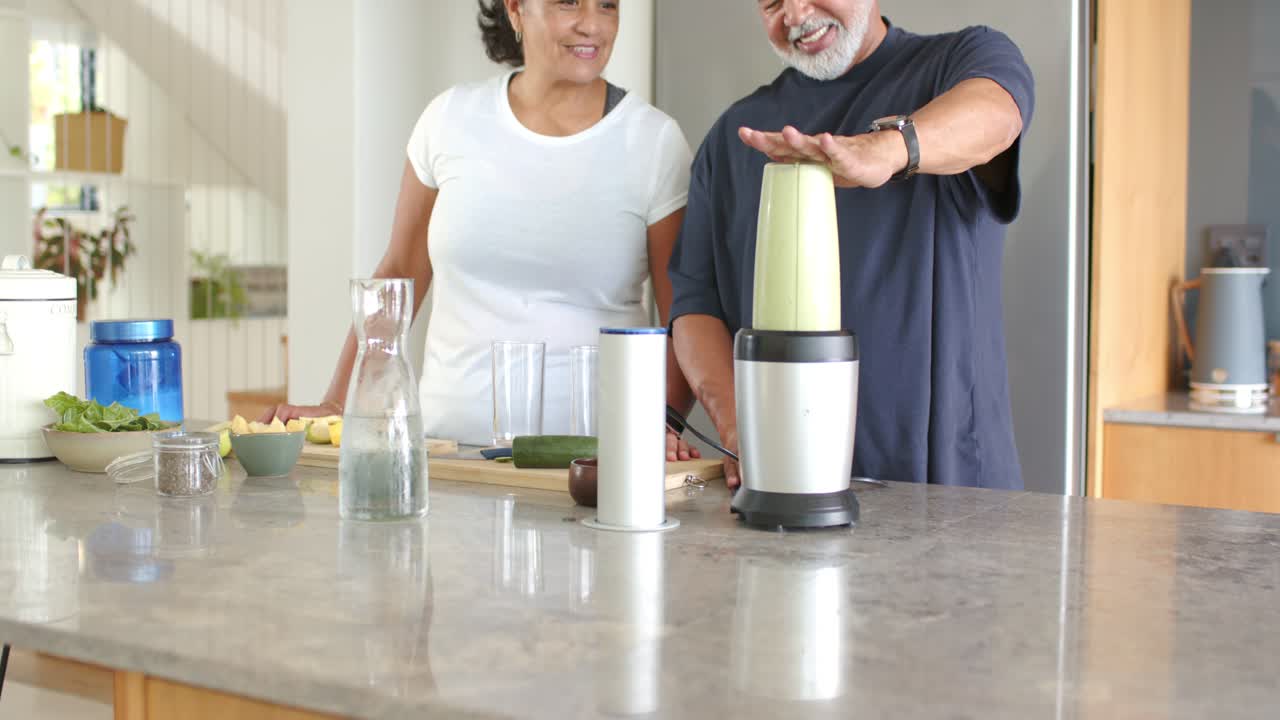 Coppia afroamericana, con l'uomo che preme il coperchio del frullatore e la donna che osserva, mentre preparano una bevanda verde al bancone della cucina.