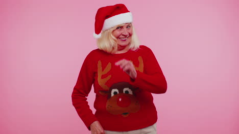 Grandmother-woman-in-Christmas-sweater-with-deers-waves-hand-palm-in-hello-gesture-welcomes-someone