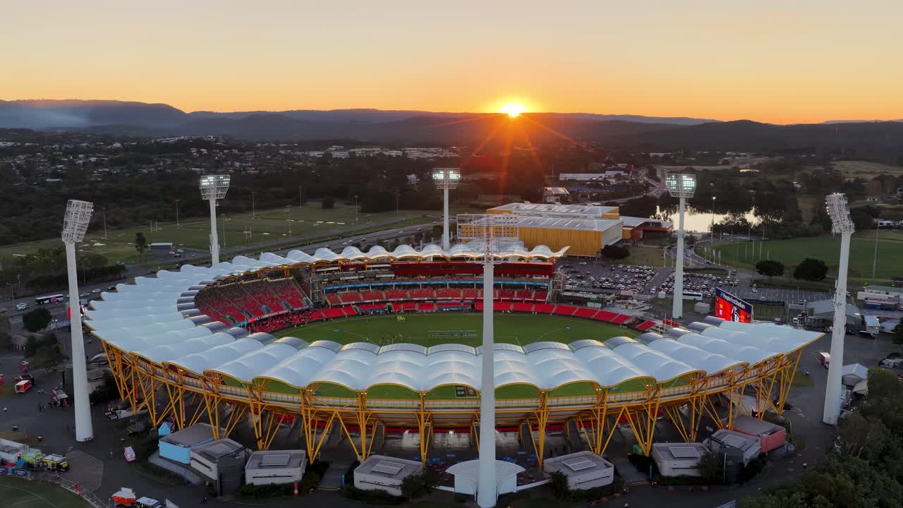 Drone footage circles a modern sports stadium at sunset, capturing illuminated field lights, surrounding landscape, and gentle river reflections under warm golden hour lighting