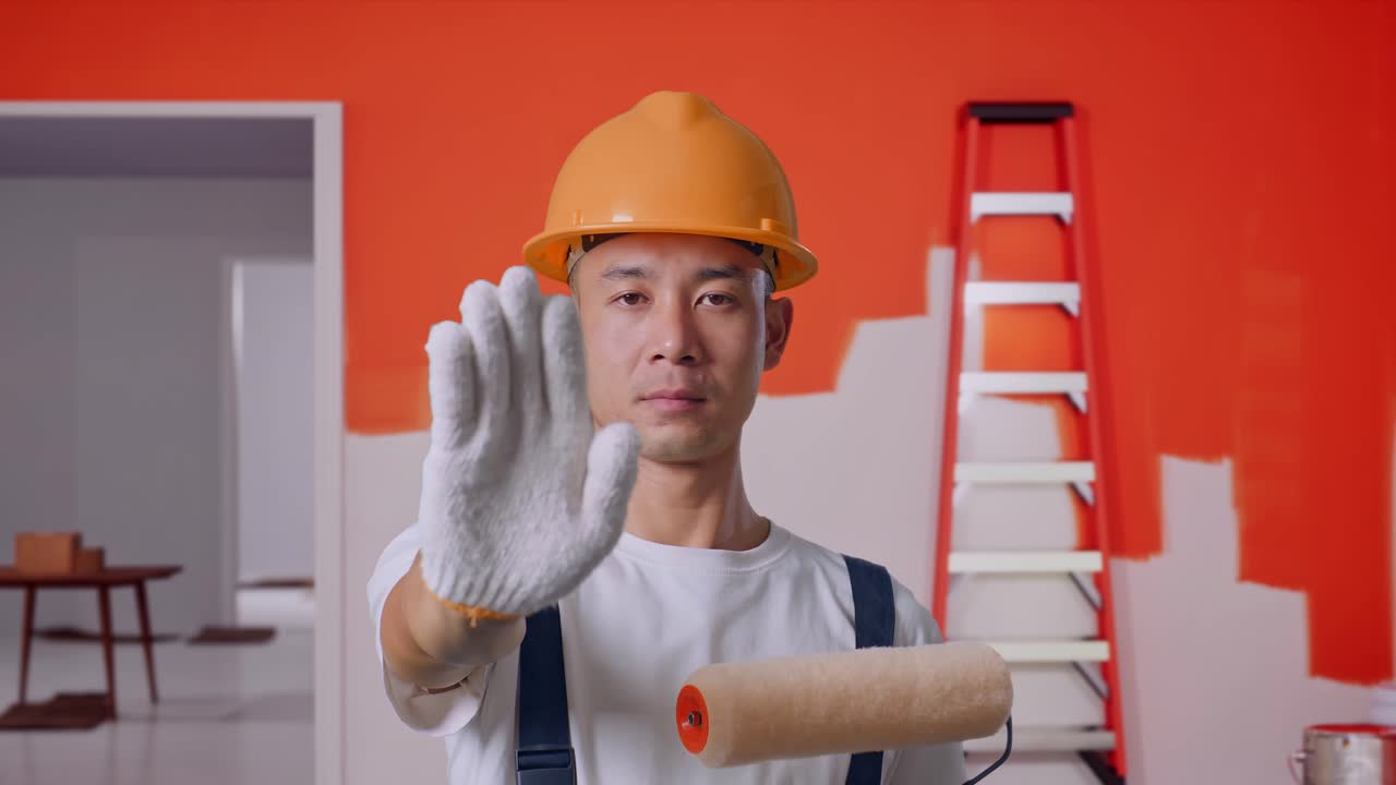 Close Up Of Asian Man Painter Wearing Safety Helmet Looking At Camera And Disapproving With No Hand Sign While Standing With Painting Wall In Room