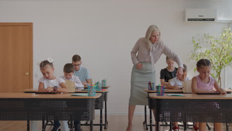 Female-teacher-controlling-learning-process-in-primary-school.-A-female-teacher-walks-between-the-desks-and-looks-after-the-completion-of-the-task