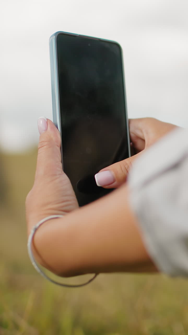 vista de cerca de la mano de la dama de manga larga tomando una foto de alguien en el fondo, la hierba alta la rodea mientras se centra en capturar el momento, con un paisaje rural suavemente borroso