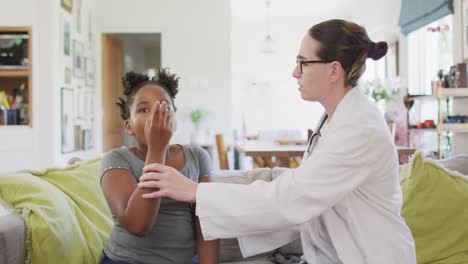 African-american-girl-and-caucasian-female-doctor-sitting-on-sofa-in-living-room-using-inhaler