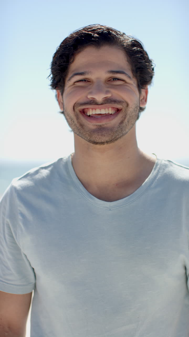 Vertical video: Smiling man enjoying sunny day at beach, wearing casual t-shirt