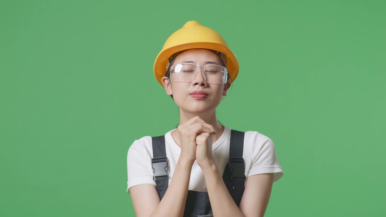 緑色の画面の背景のスタジオに立って何かのために祈っている保護眼鏡と安全ヘルメットを着たアジアの女性労働者のクローズアップ