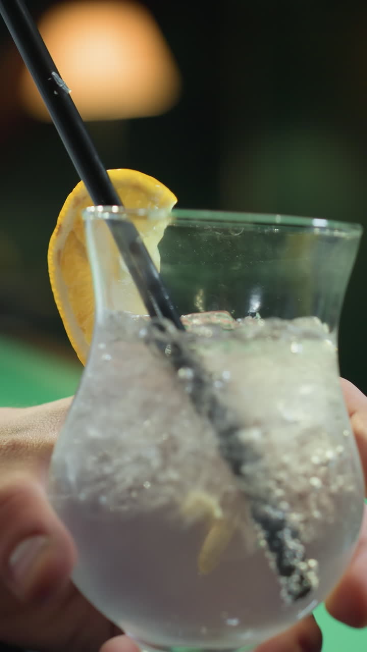 primer plano de una persona, cara no visible, con camisa blanca y pantalones grises, sosteniendo un vaso de bebida de limón con paja negra. los cubos de hielo y las rebanadas de limón agregan detalles refrescantes a medida que la cámara hace zoom
