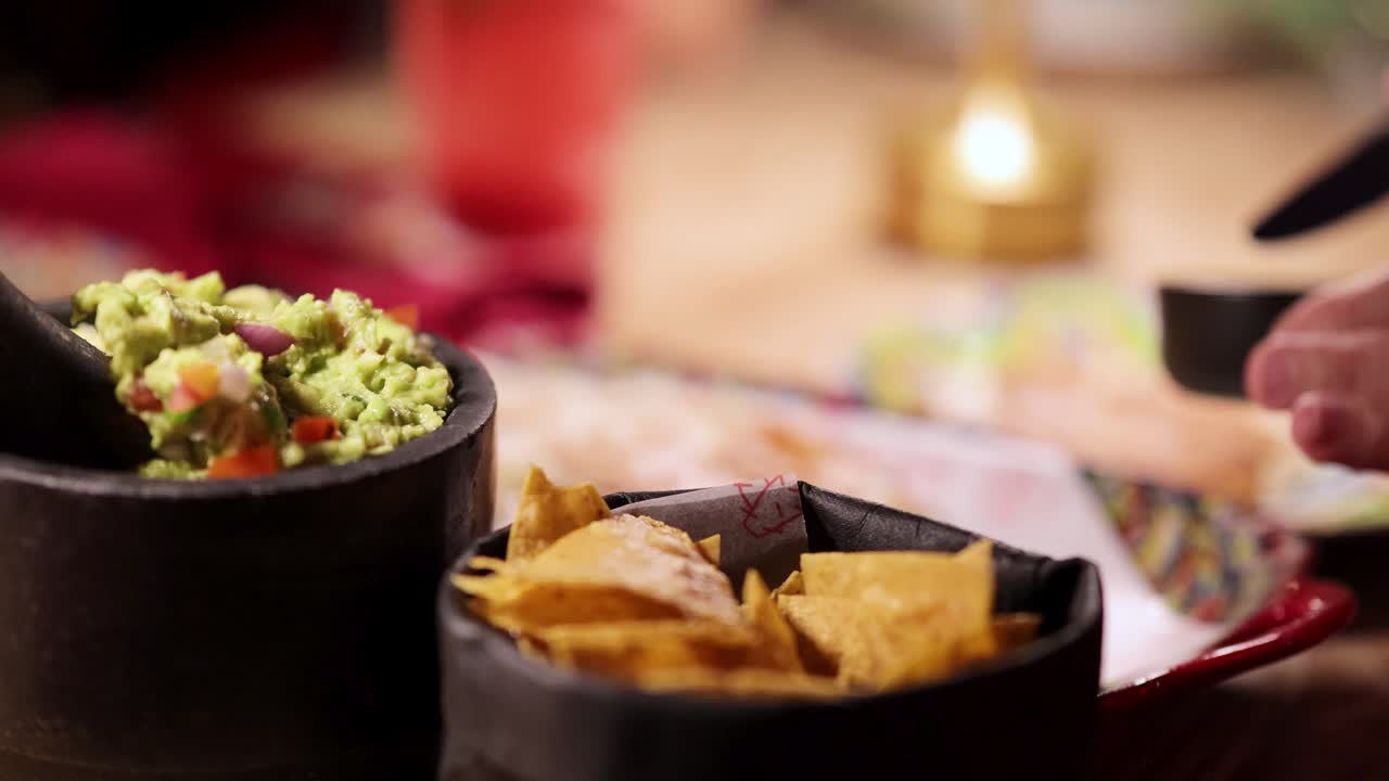 A hand reaches for tortilla chips with guacamole in a cozy, warmly lit restaurant setting