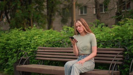 woman sitting on wooden bench outdoors, deep in thought after a phone call, resting left hand on her leg, surrounded by vibrant greenery, trees, and building in background