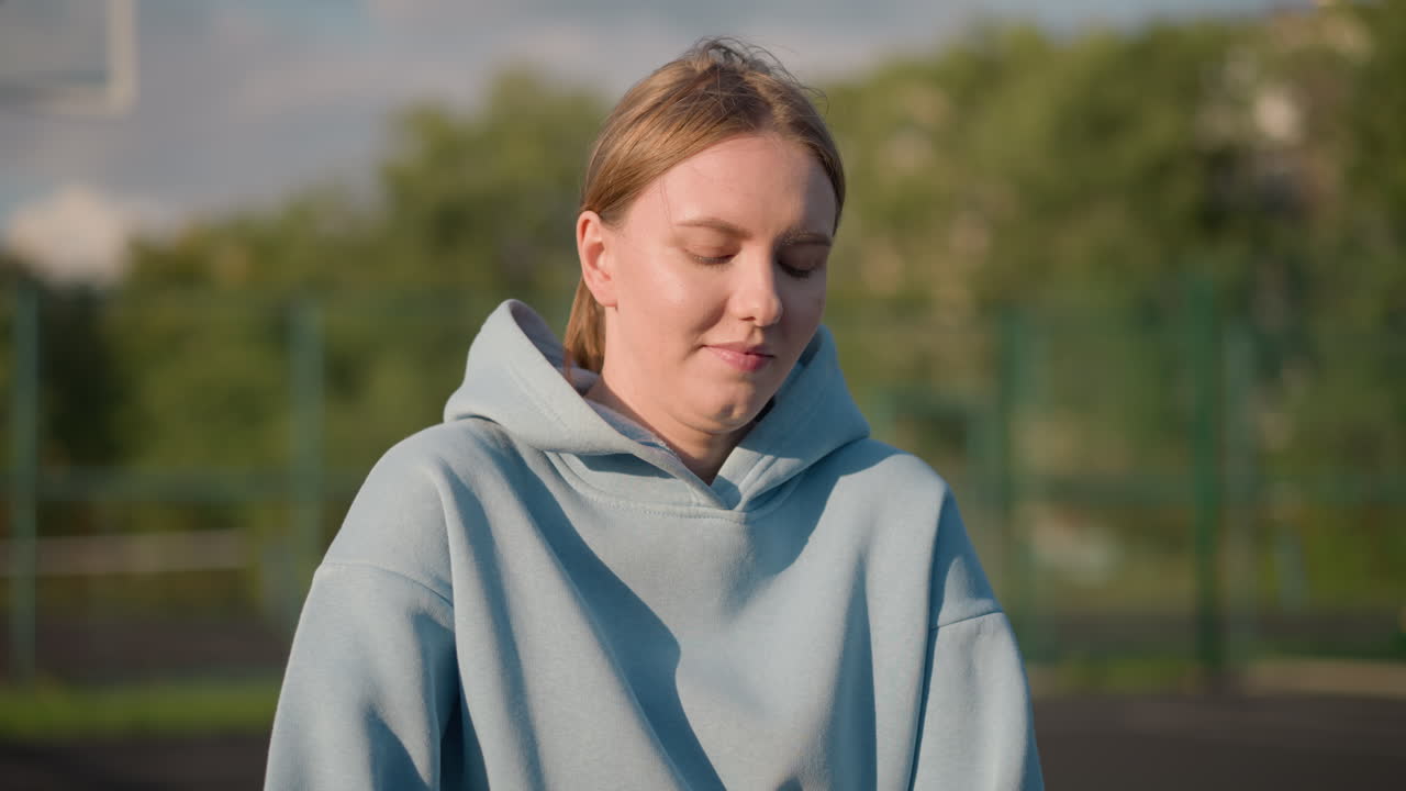 молодая леди занимается фитнес-упражнениями, стоя на коленях вперед, волосы привязаны назад, с размытым фоном с деревом и неясными фигурами на расстоянии