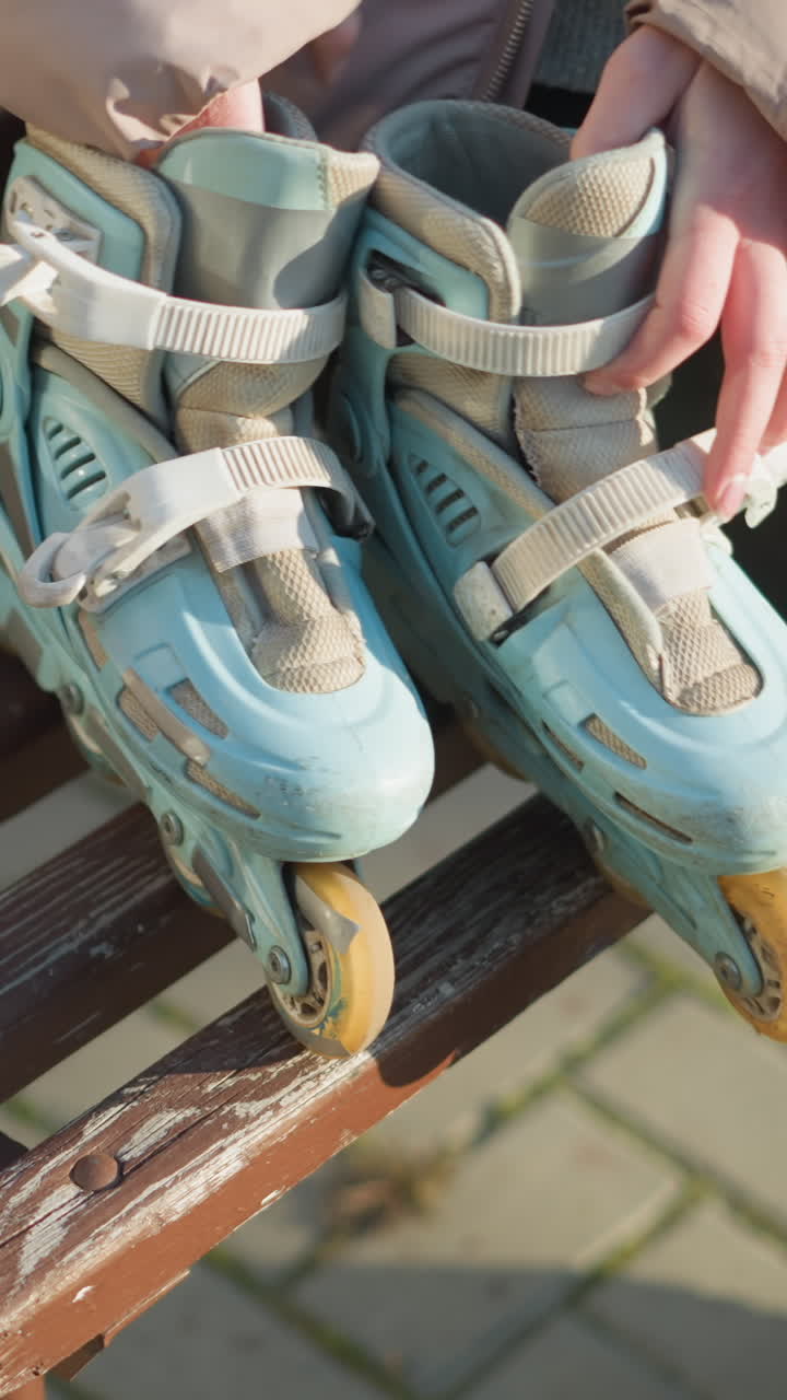 Close-up of a person holding rollerblades with both hands while sitting down on a park bench. The wooden bench is visible with signs of wear, and the person's rollerblades are placed beside them