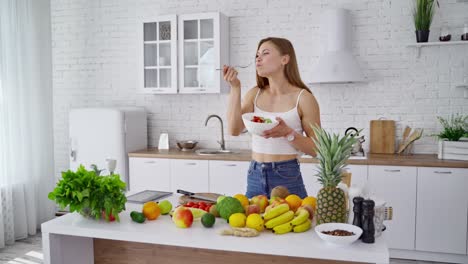Young woman in the kitchen. Beautiful girl standing near the table with organic food. Lovely female enjoys eating fresh salad on the modern kitchen background. Dieting concept.
