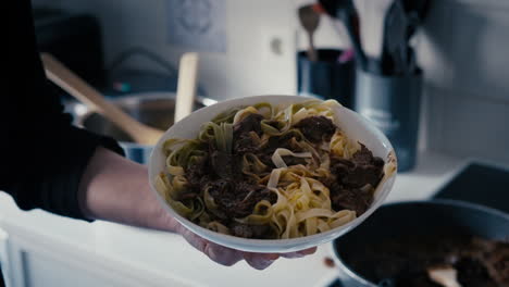 close up of a hand revealing tagliatelle pasta plate with meat sauce in the white kitchen with pasta and meat in the background