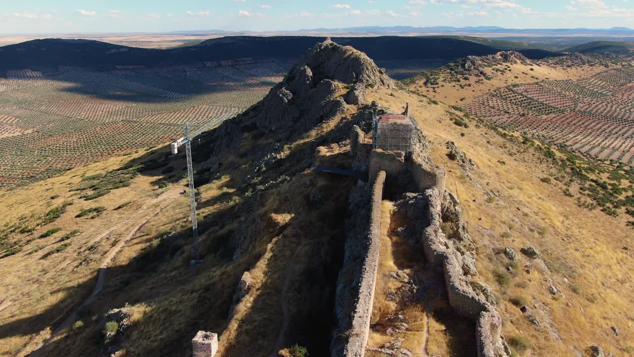 Un drone in discesa con inclinazione verso l'alto rivela la forma allungata, simile a una barca, del Castello di Peñas Negras, arroccato su una stretta cresta rocciosa con lavori di restauro visibili