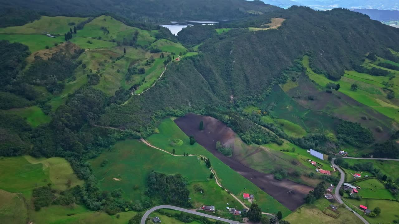 Drone pullback above the green valley near Laguna de Pantano Redondo, showing hills, fields, forests and rural roads