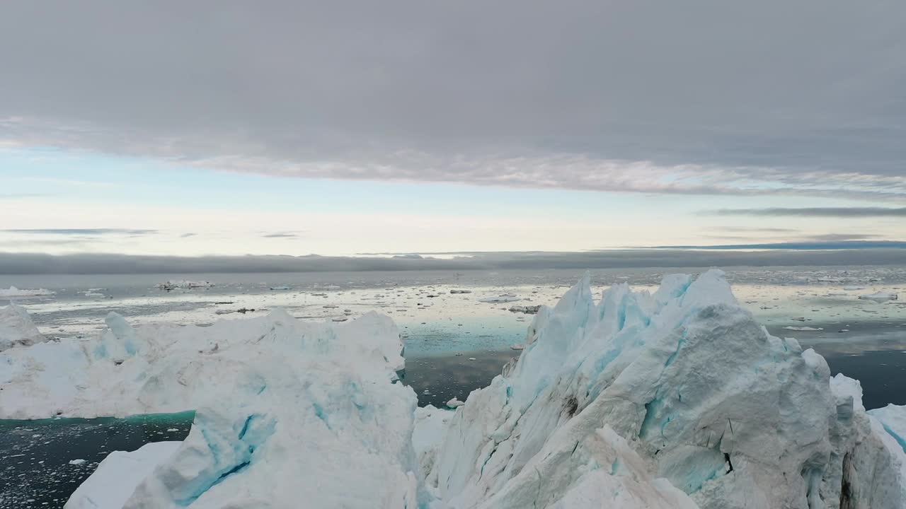 Aerial drone view of tall icebergs floating in calm Arctic waters under a cloudy sky in Greenland, showing the stunning beauty and vastness of the frozen polar environment