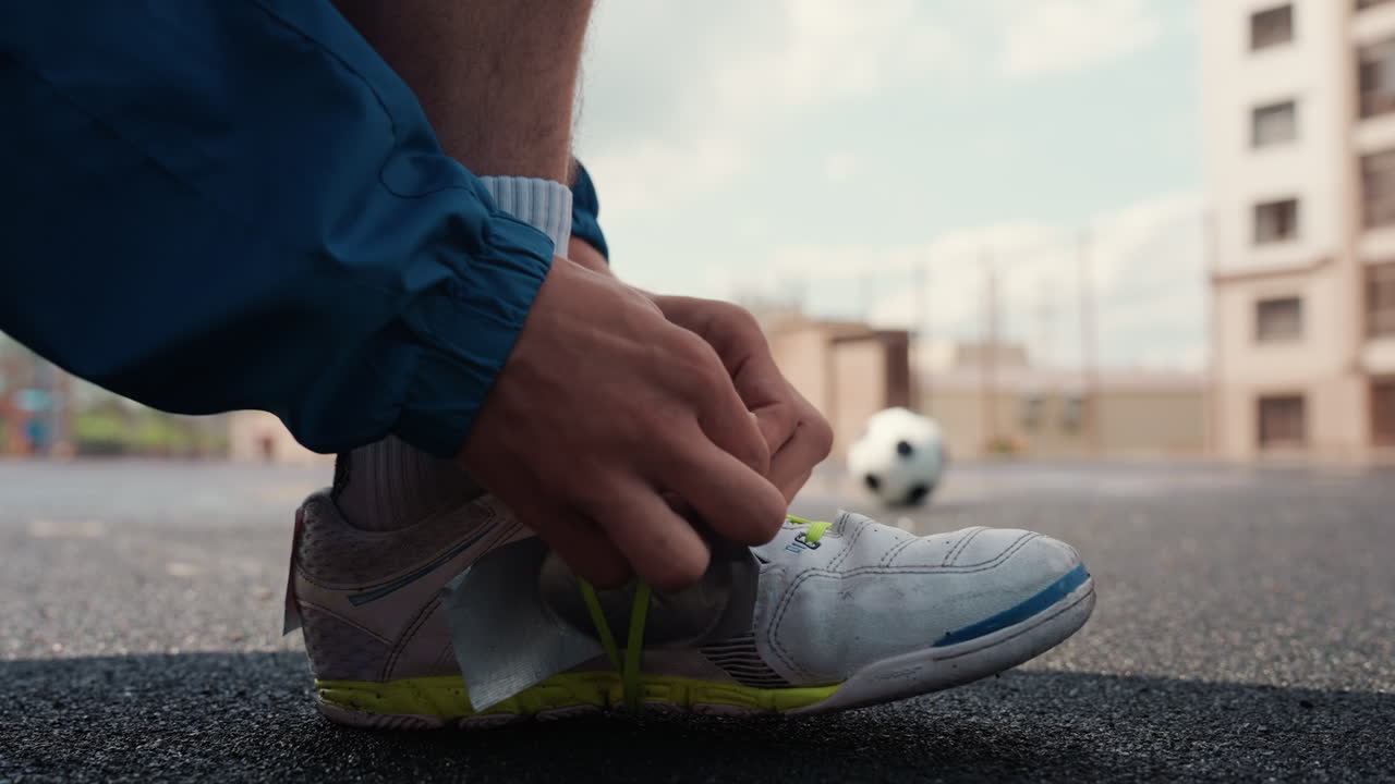 Primo piano di un atleta che si allaccia la scarpa da ginnastica bagnata accanto a un pallone da calcio su un campo di asfalto, gocce di pioggia e cielo nuvoloso, mani concentrate sull'allacciatura, rituale pre-partita, atmosfera urbana grintosa e umore determinato