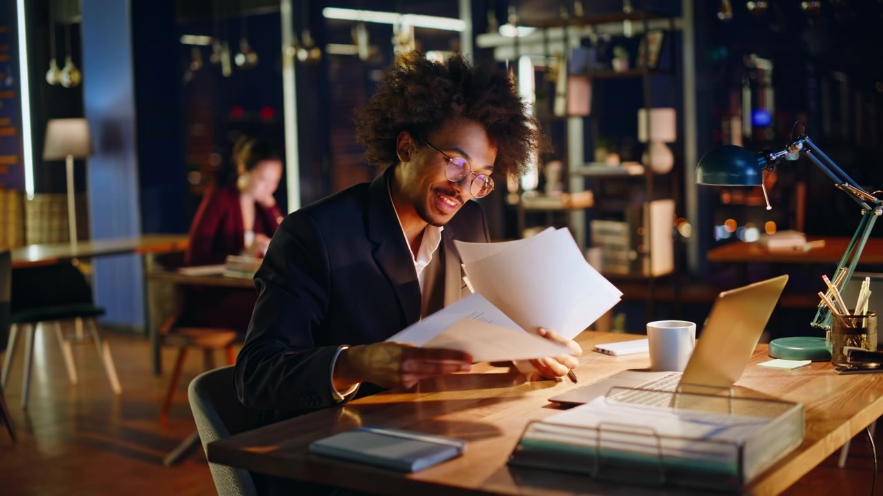 Young specialist gesturing video call night office. Man reading paper excited.