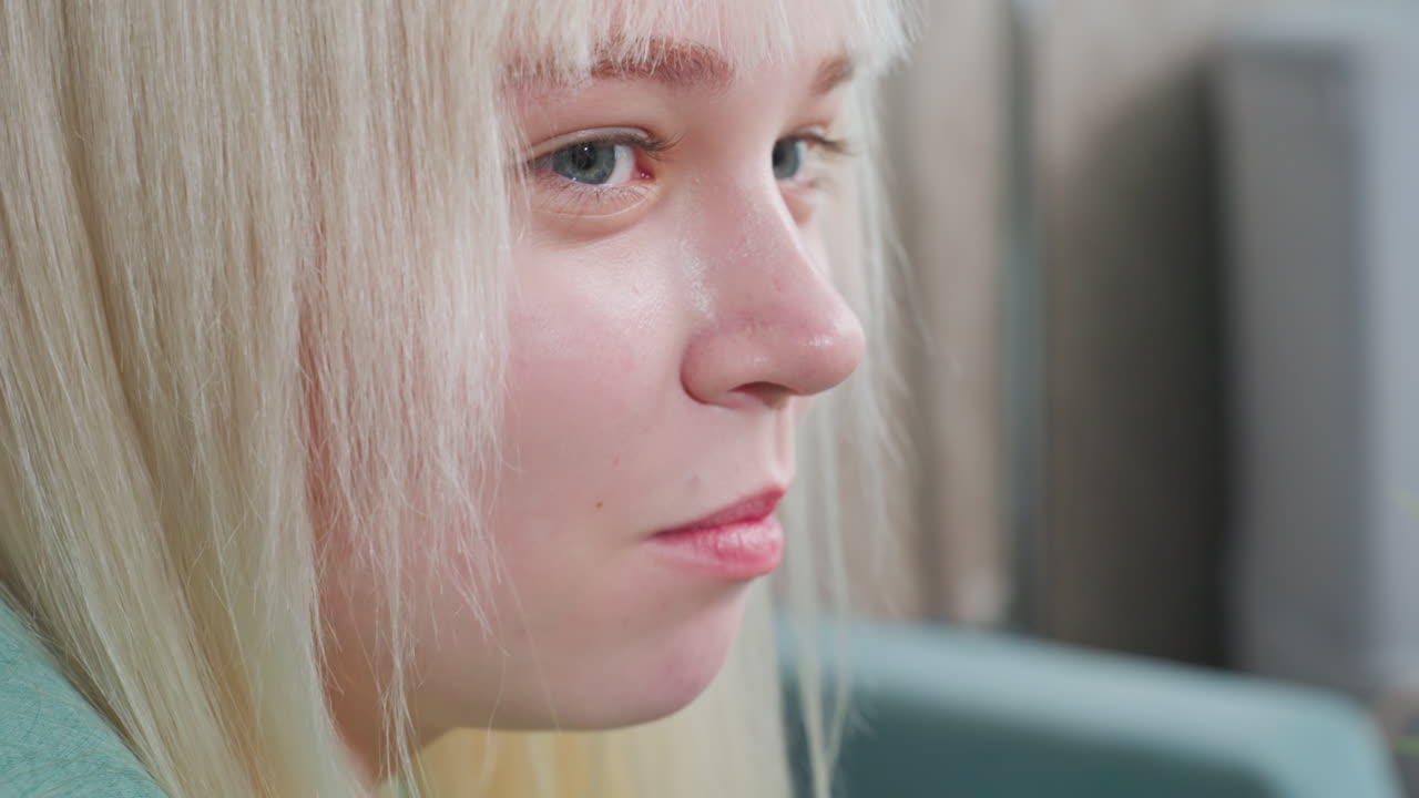 Close up of young blonde woman in green shirt enjoying popcorn while talking to someone in cozy sitting room with soft lighting and textured wallpaper background
