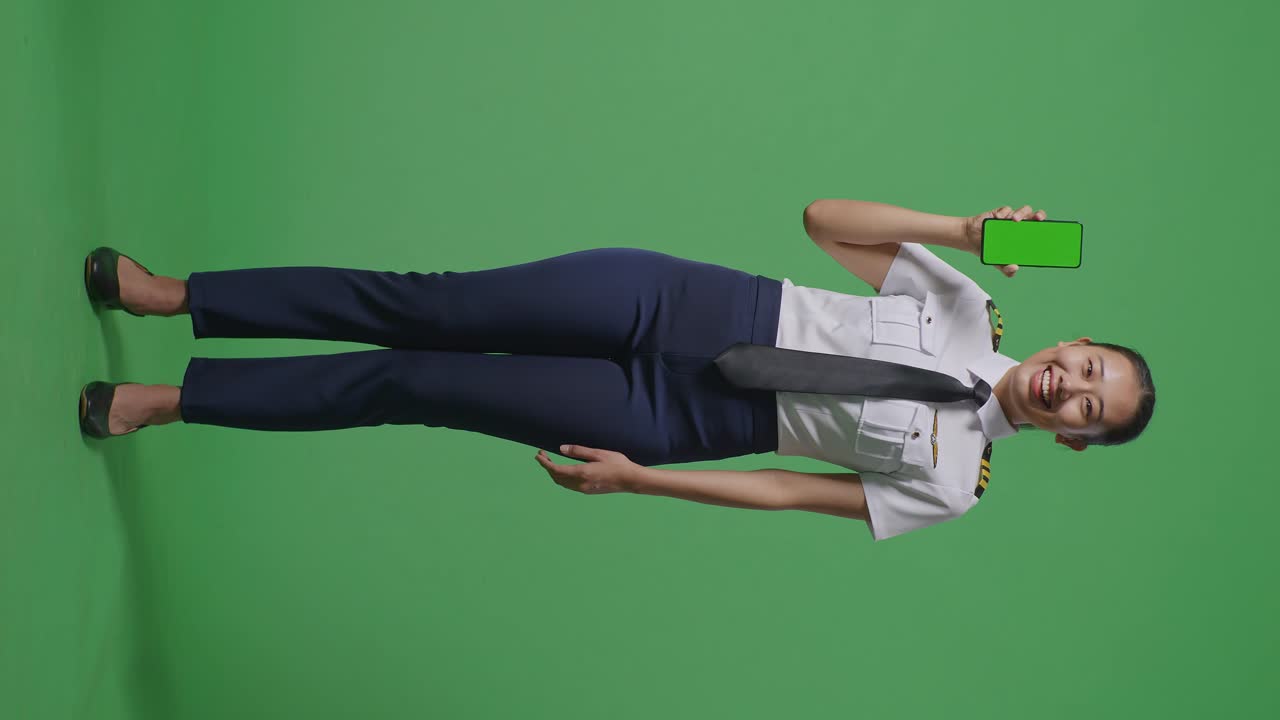 아시아 여성 조종사가 미소 짓고 녹색 화면 배경 스튜디오에 서 있는 동안 카메라에 초록 화면 스마트폰을 보여주고 있습니다.