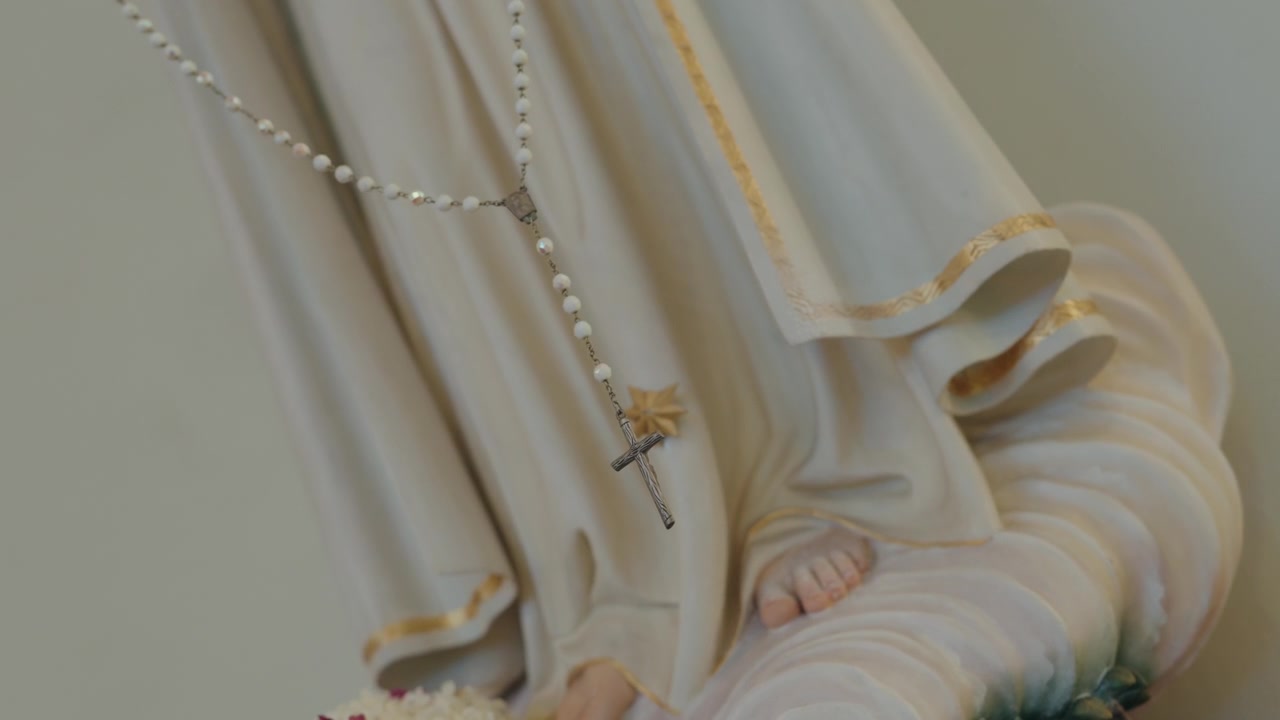 Cuentas de rosario blancas y crucifijo de plata colgando sobre el manto de la estatua de Nuestra Señora.